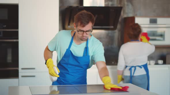 Young Man with Detergent Spray and Cloth Cleaning Cooking Top When Woman Wiping Furniture in Kitchen