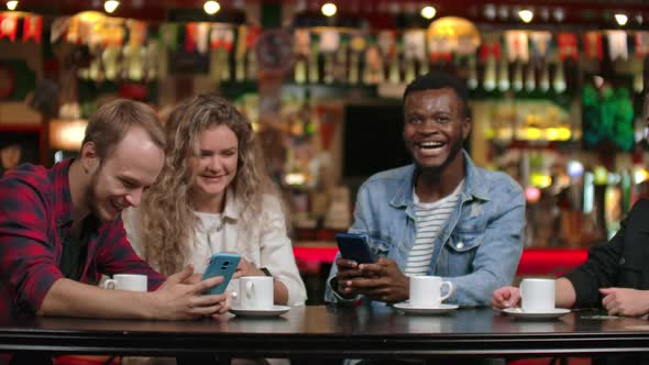 Four Friends Sitting in a Cafe Laugh and Smile While Looking at the Screens of Phones and Exchanging