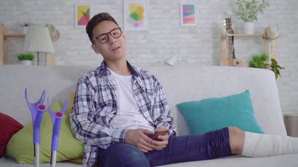 Cheerful Asian Young Man with a Broken Leg in Bandages Uses a Smartphone Looking at Camera and