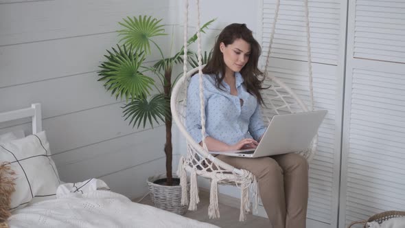 Girl Sitting with a Laptop on Her Lap in a Cozy Hanging Chair. Young Female Freelancer Working