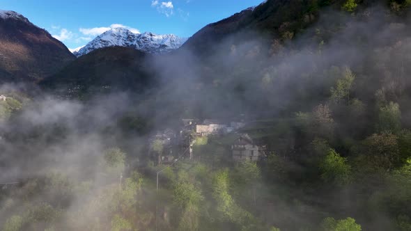 Flight through fog toward picturesque Italian village in alp mountainside; drone