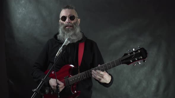 Bearded hairy rockstar in retro round sunglasses plays on red electric guitar with black background