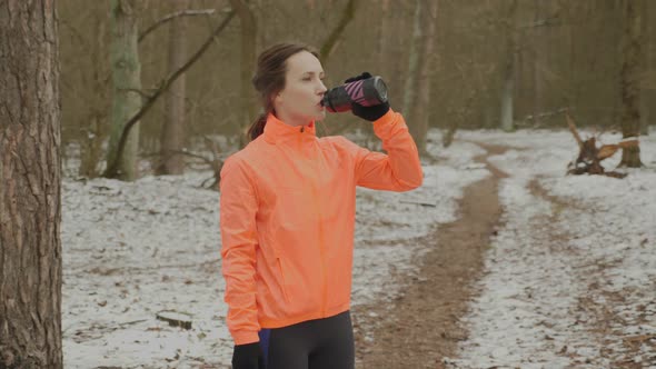Young woman drinking water after running training in cold snowy winter park wearing orange jacket