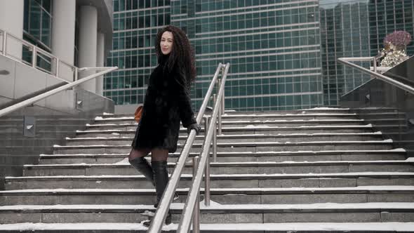 A Curlyhaired Stylish Middleaged Brunette Woman in a Black Fur Coat Walks Alone in the City on a