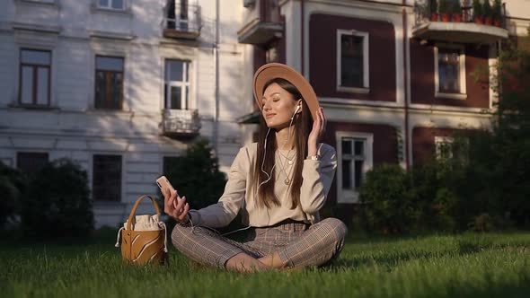 Young Slender Satisfied Woman in Modish Hat Sitting on Green Grass Near Cozy Houses