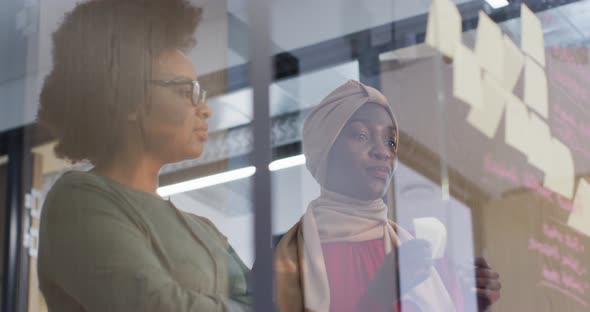 Two smiling african american businesswomen brainstorming using memo notes on glass wall in office