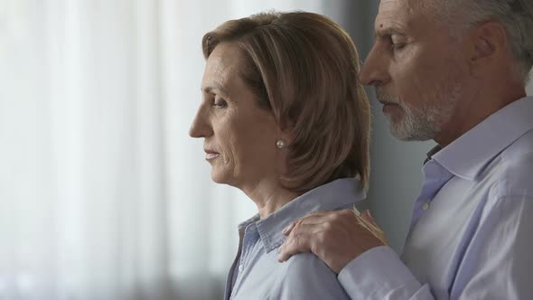 Sad Elderly Female Standing by Window, Male Taking Her by Shoulders, Support