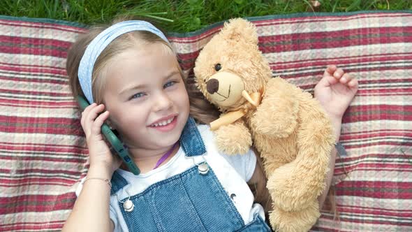 Pretty Child Girl Laying on Green Lawn with Her Teddy Bear Talking on Mobile Phone