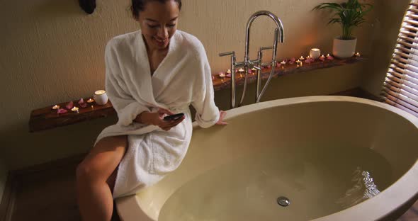 Happy biracial woman with vitiligo sitting in bathrobe, running bath and using smartphone
