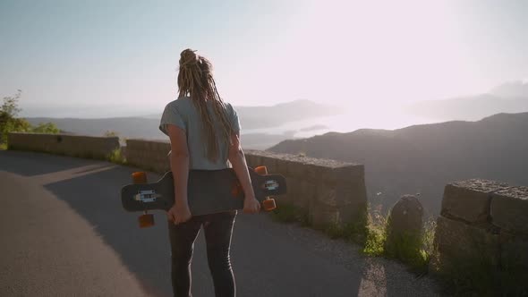 Camera following dreadlocks woman walking with longboard on mountain background