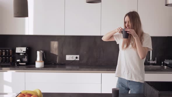 A Young Woman is Dancing in the Kitchen and Drinking Coffee