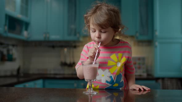 Cute little girl in pink t-shirt drinking berry smoothie with paper straw at home