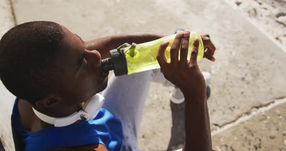 African american man sitting and drinking from water bottle taking break in exercise outdoors