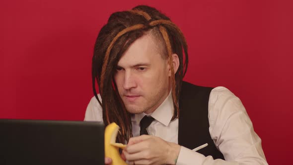 Young man works on laptop and eats banana on red background.