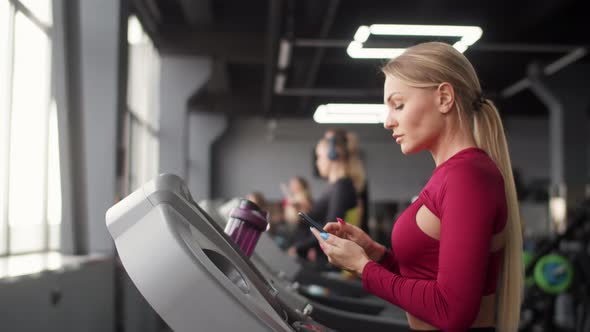 Beautiful young blonde woman in red sportswear using smart phone during workout on treadmill at fitn