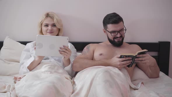 Portrait of Happy Caucasian Spouses Lying in Bed in the Evening. Cute Blond Woman Using Tablet, Man