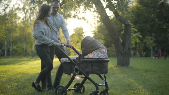 Side View Wide Shot of Cheerful Young Couple of Parents Walking with Baby Carriage in Sunlight and