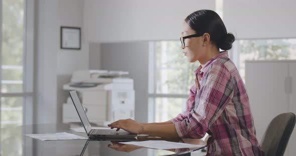 Side View of Asian Businesswoman Working on Laptop Sitting at Desk in Modern Office