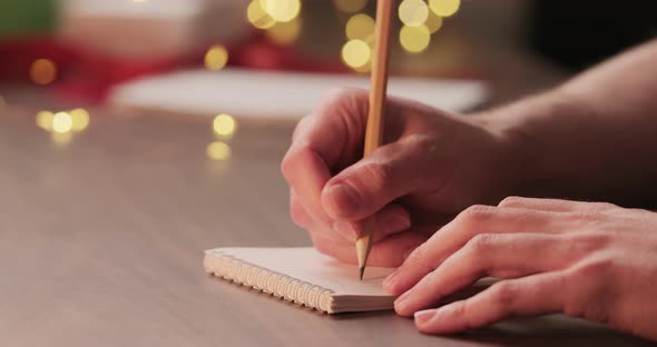 Man Writes Shopping List for Holidays in Notepad with a Pencil Under Warm Light in the Evening