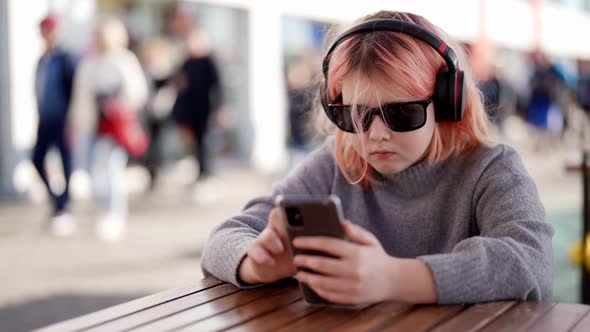 Stylish Teenage Girl with Pink Hair Listening to Music with Headphones Outdoors in a Cafe at a Table