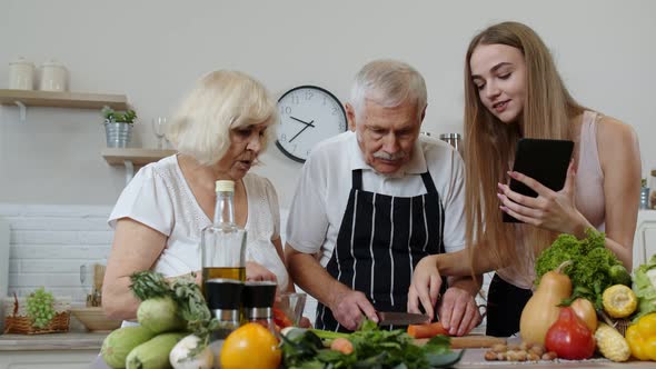 Senior Grandparents Couple Cutting Vegetables for Salad, Listening Recipe From Girl with Tablet