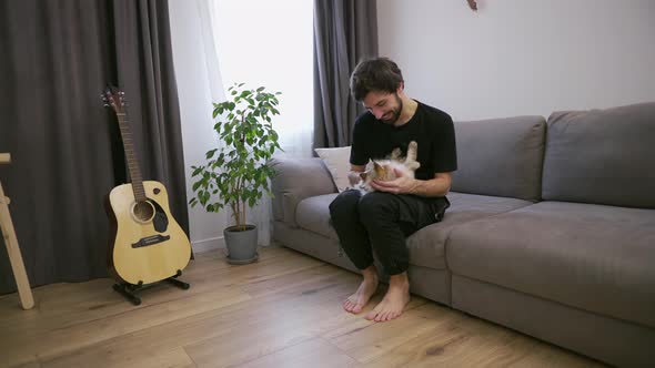 Young Man Sitting on a Sofa Caresses a Tabby Cat on Knees