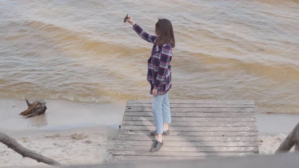 Wide Shot of Attractive Caucasian Girl Taking Selfie Standing on Pier. Brunette Young Woman Posing