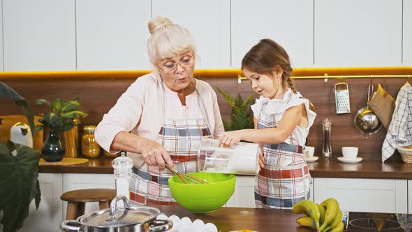 Grandmother Whipping Eggs By Whisk in Bowl and Her Little Granddaughter is Adding Flour They Going