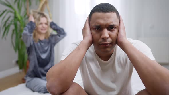 Frustrated African American Young Man with Hands Covering Ears Sitting on Bed Looking at Camera As