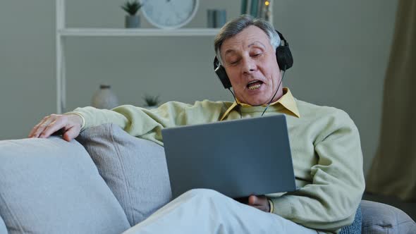Excited Happy Aged Man Watching Movie Using Laptop and Headphones Sitting Lying on Comfy Couch