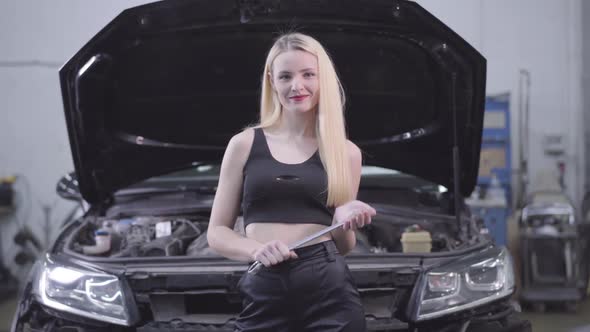 Sexy Blond Caucasian Woman Standing at the Background of Open Car Hood with Adjustable Wrench 