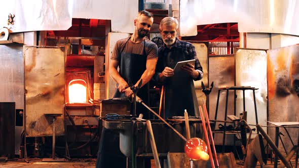 Glassblowers discussing over digital tablet while working on a molten glass