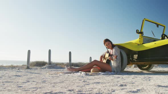 Happy caucasian woman sitting beside beach buggy by the sea playing guitar
