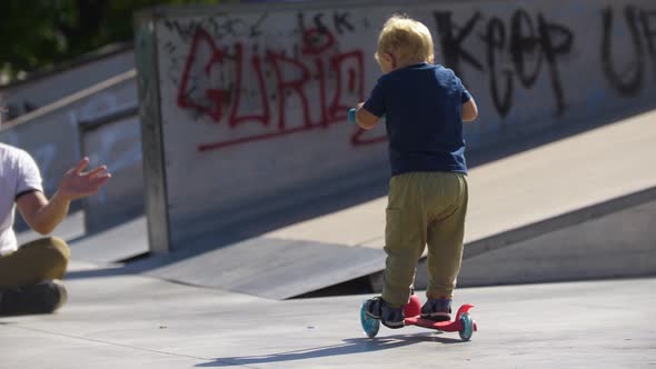 Little Blonde Boy Skating on the Scooter in the Skate Park and His Father Watching Him