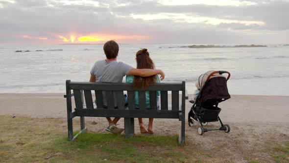 Rear View of Loving Family Couple Sitiing By a Ocean Looking at Sunset with Baby Stroller