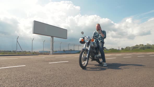 Young Man Motorcyclist with Smart Phone in Hands on Custom Motorcycle on Street