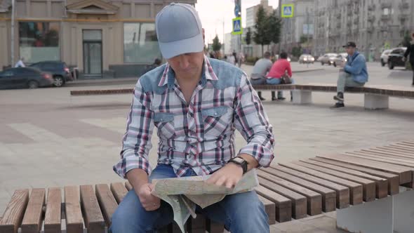 Caucasian Man Sitting on a Bench in a Square in the City Studying a Map of City