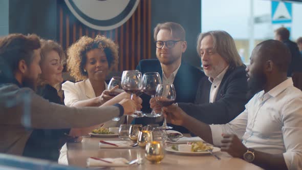 Group of Happy Friends Meeting and Having Dinner Clinking Wine Glasses in Modern Restaurant