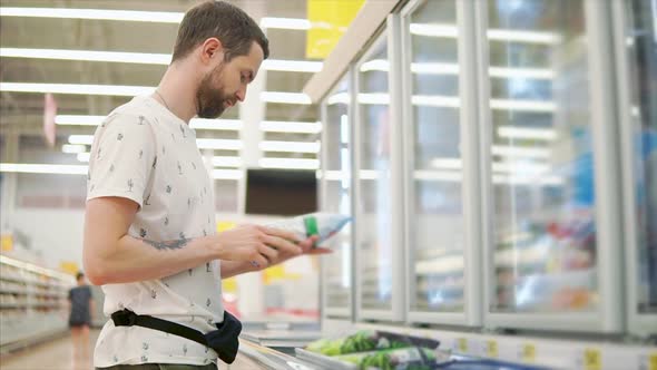 Young Man is Examining Pack with Frozen Vegetables in a Hall of Supermarket