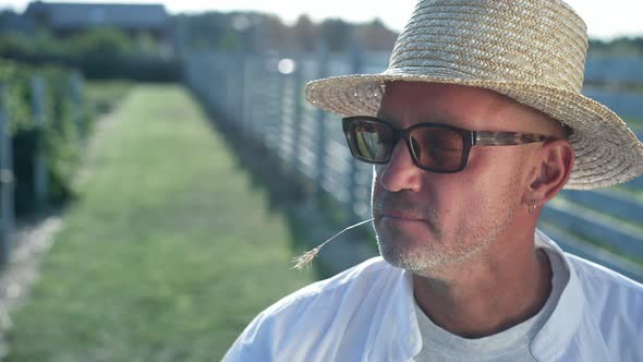 Closeup Confident Farmer in Straw Hat in Sunbeam on the Right with Green Garden at Background