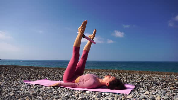 A Young Woman with Black Hair Doing Pilates with the Ring on the Yoga Mat Near the Sea on the Pebble