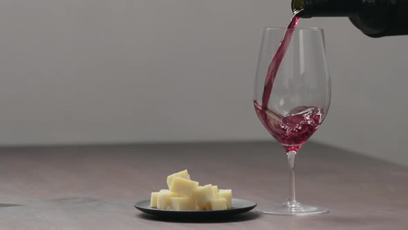 Slow Motion Pour Red Wine Into Wineglass with Hard Cheese on Background on Wood Table