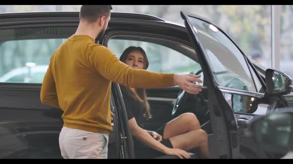 Young Caucasian Man in Mustard Sweater Opening Car Door and Talking To Beautiful Girl Sitting in Car