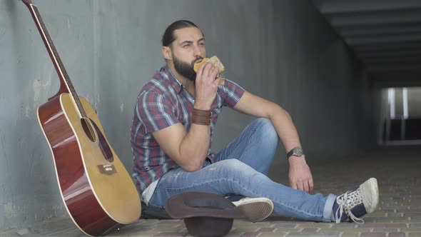 Poor Caucasian Man Sitting with Guitar and Hat in Urban Underground Crossing and Eating Bun. Wide