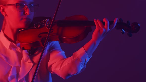 Woman Violinist in Glasses and a White Blouse Plays the Violin Standing in Studio Illuminated By a