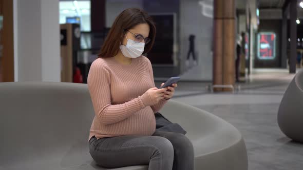 A Young Pregnant Woman in Glasses and a Medical Mask Sits with a Phone on a Bench