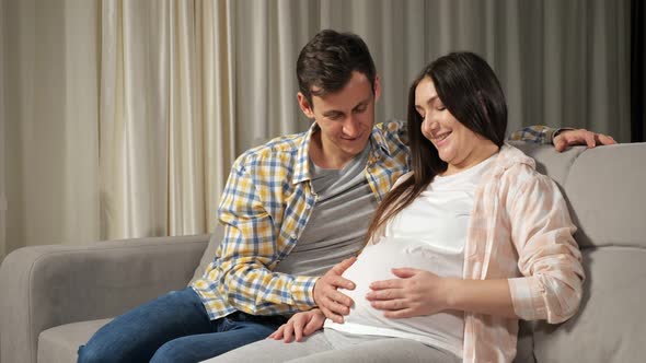 Husband Caresses Pregnant Stomach of Wife Sitting on Sofa