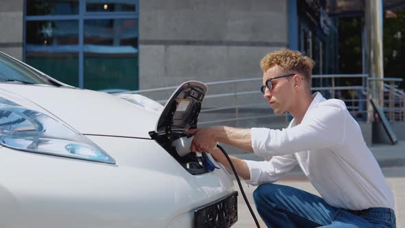 A Young Curly Man Connects an Electric Car to the Charger