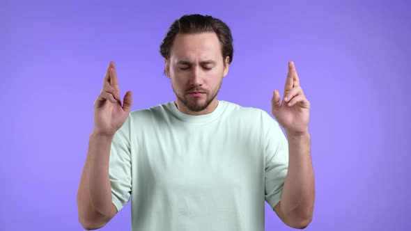 Handsome Man Praying with Crossed Fingers Over Violet Background