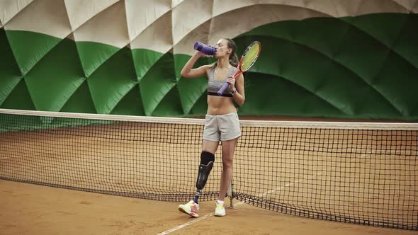 Seductive Tennis Girl with the Leg Prosthesis is Standing Near the Net on the Tennis Court Drinking